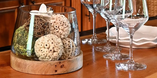 Styled glass hurricane vase on wooden table with wine glasses.